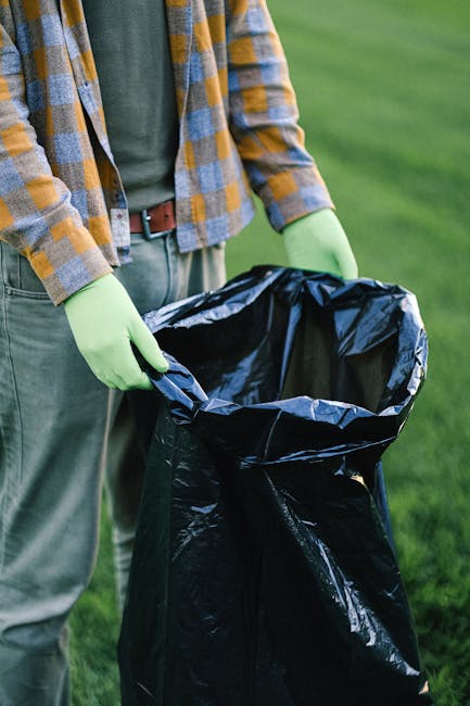 A sanitation worker, wearing a high-visibility yellow and orange vest, is seen standing on a paved road next to a large red rubbish collection truck, which is actively engaged in waste collection. The