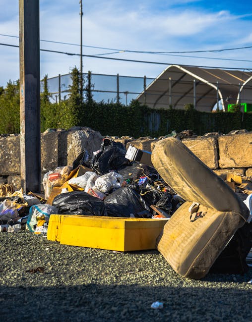 A close-up view of a pile of mixed waste and rubbish situated on a gravel surface next to a low stone wall outside, with a yellow plastic container and a discarded used car tire partially obstructing 