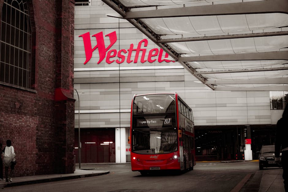A red double-decker bus with a modern design is positioned centrally in the image, approaching a bus stop outside a large retail store named Westfield, as indicated by a prominent red sign on the buil